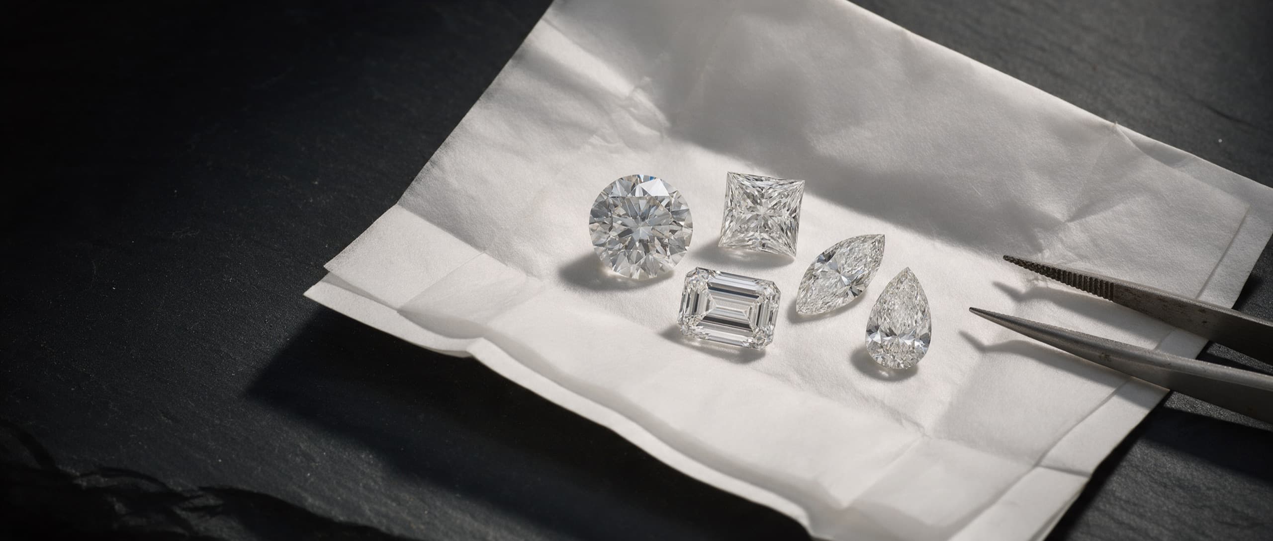 Five loose diamonds of ascending size — round, oval, emerald, cushion and pear — laid in a line on black stone