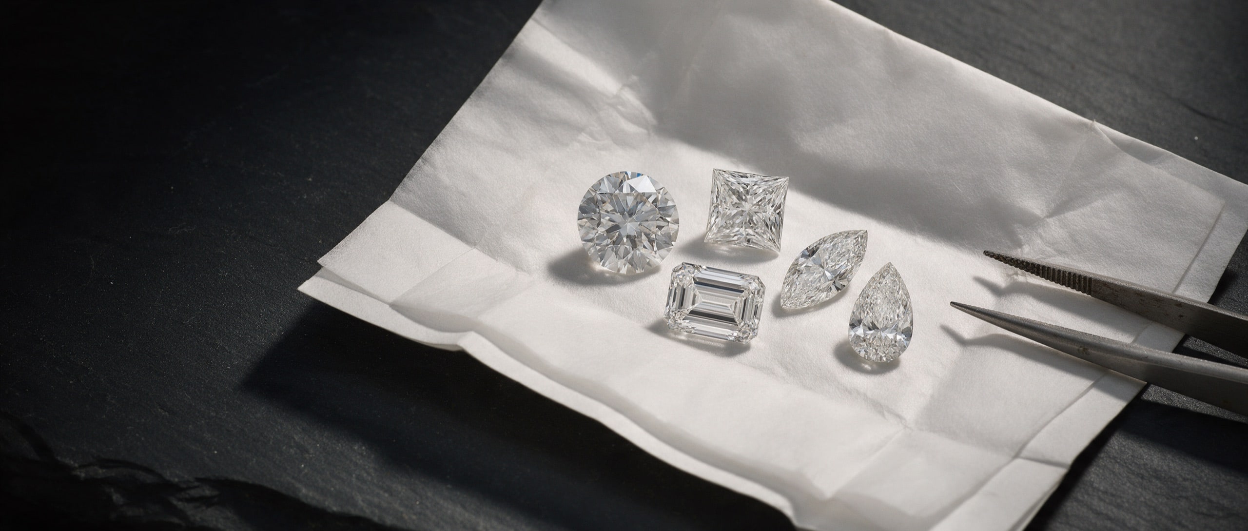 Five loose diamonds of ascending size — round, oval, emerald, cushion and pear — laid in a line on black stone