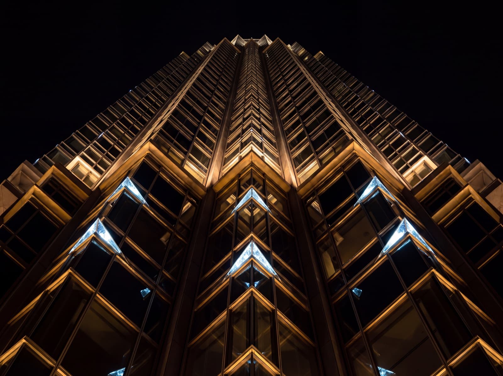 A modern glass tower at night seen from its base, facade catching warm gold interior light
