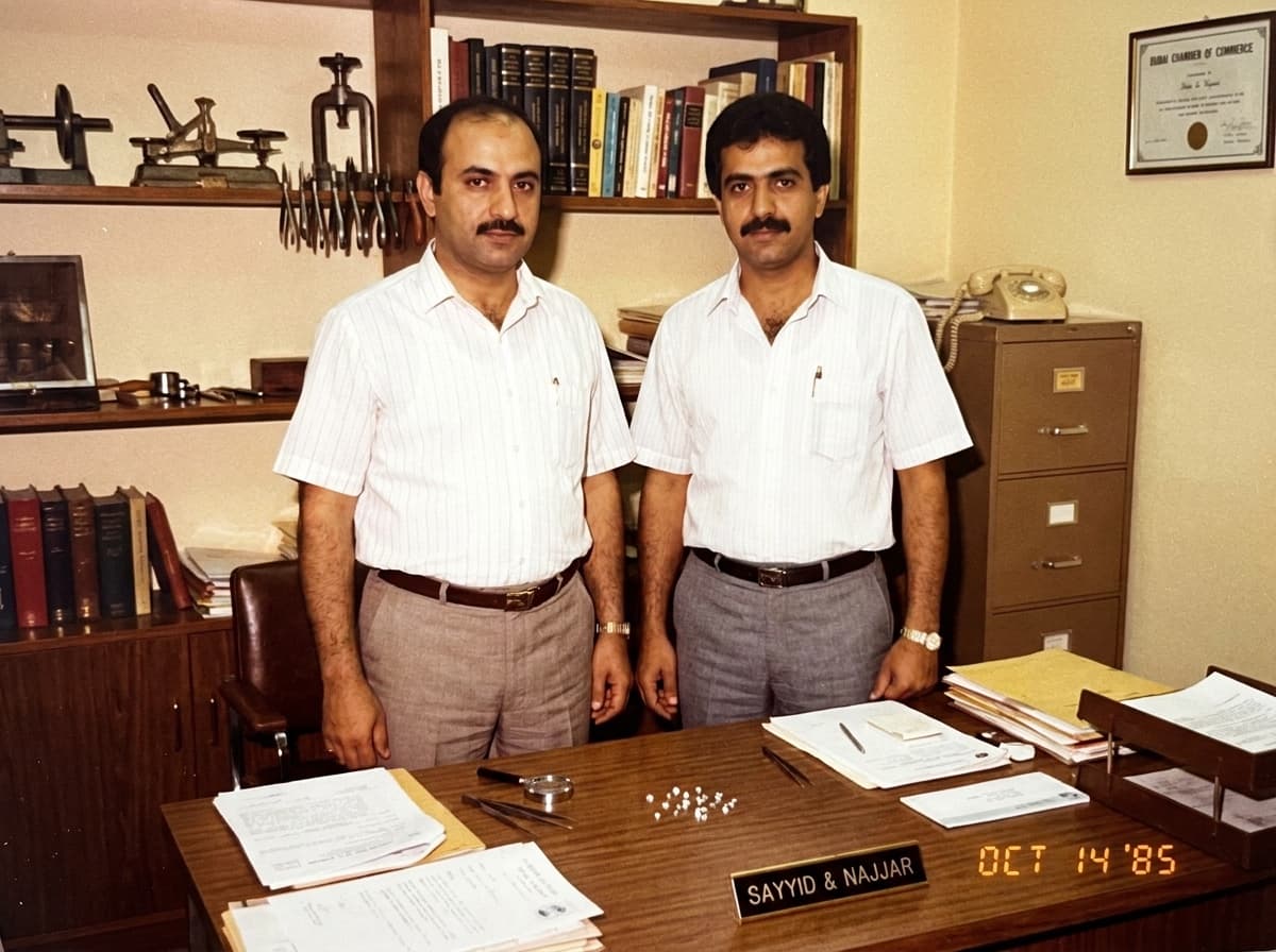 Founders Mr. Sayyid (left) and Mr. Najjar (right) standing behind their desk in the Sayyid & Najjar Co. office in Dubai in October 1985, a Sayyid & Najjar nameplate and a small parcel of loose diamonds on the desk before them